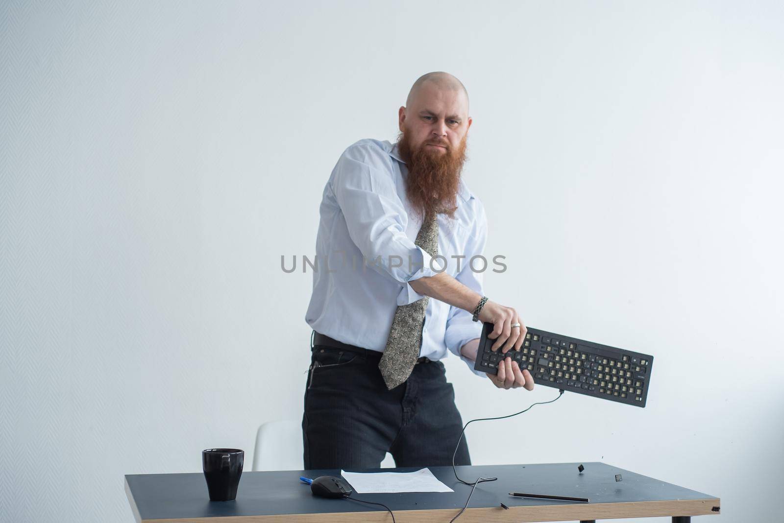 Angry bald man with a red beard in the office in a business suit crashes a computer. Manager with a nervous breakdown breaks the keyboard on the monitor. by mrwed54