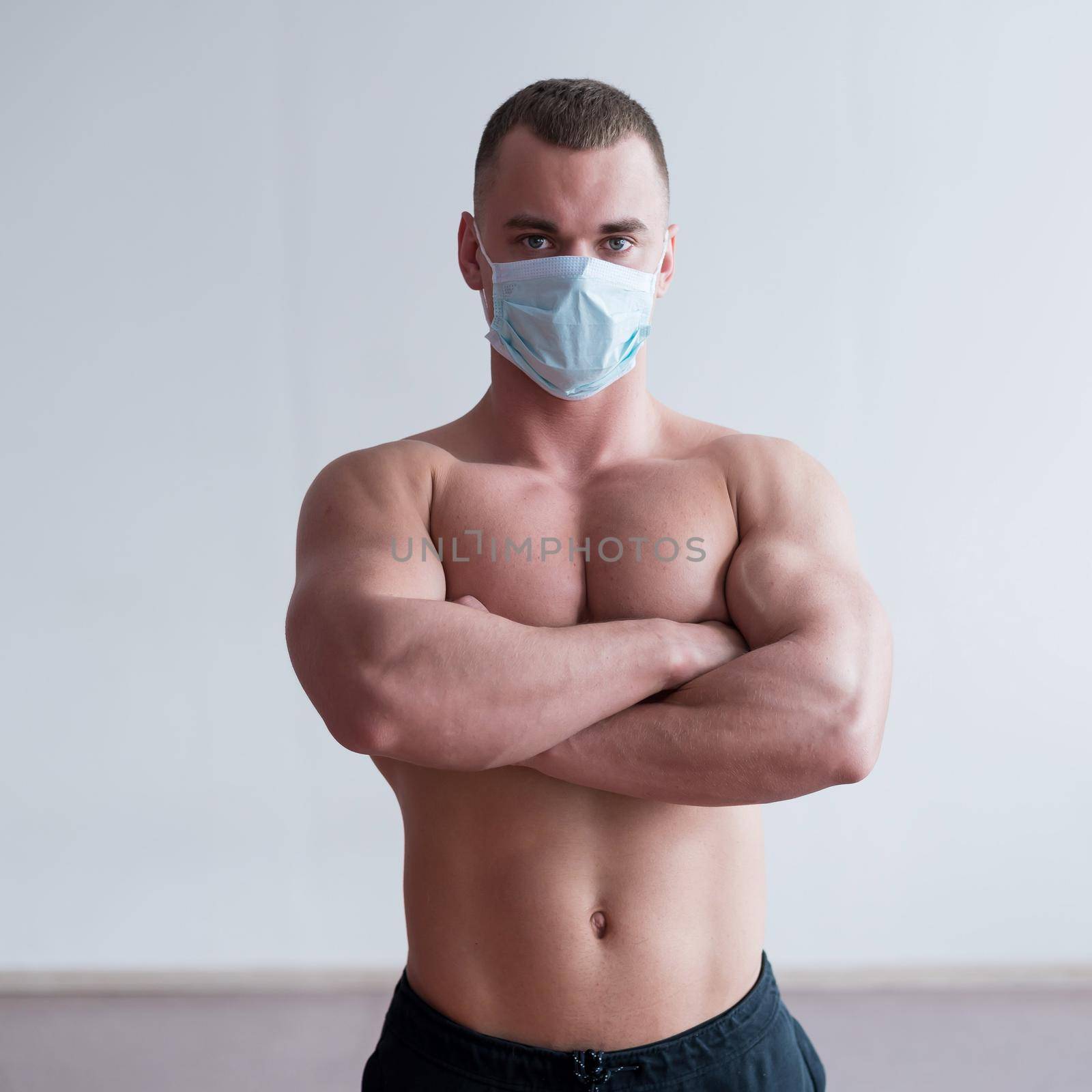 A male bodybuilder in a medical mask crossed his arms over his muscular bare chest. The guy goes in for sports in quarantine. Respiratory protection. by mrwed54