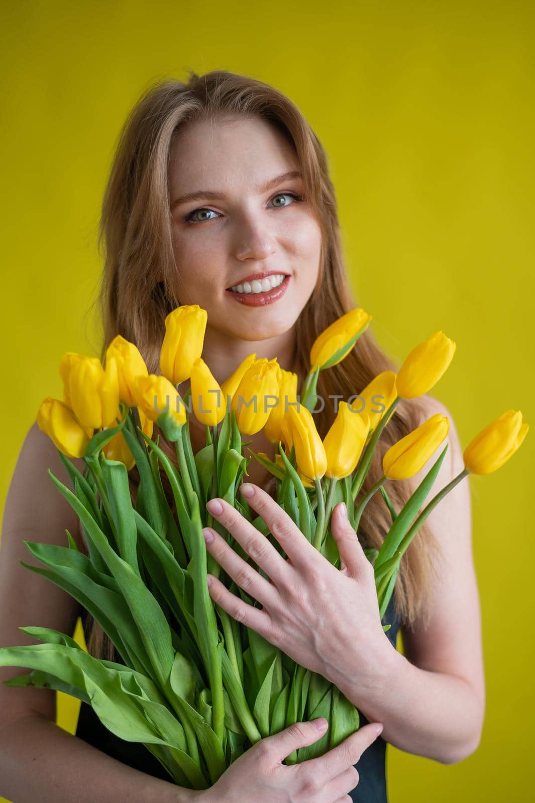 Caucasian woman with an armful of yellow tulips on a yellow background. International Women's Day. Bouquet of spring flowers.