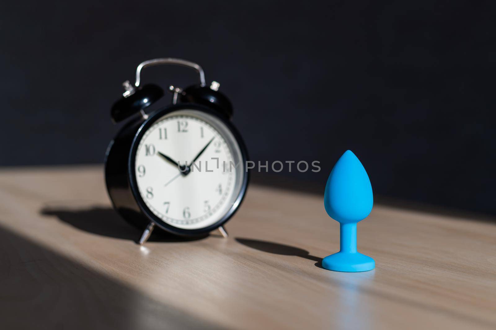 Blue latex anal plug and alarm clock on a wooden table. The time of carnal pleasures