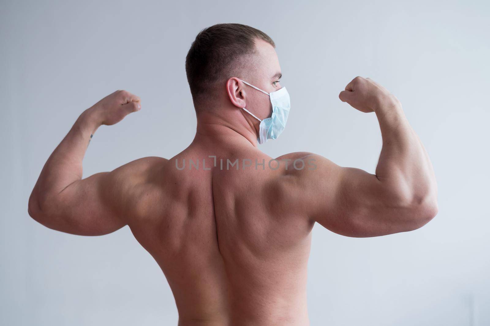 Male bodybuilder in a medical mask stands with his back. The guy goes in for sports in quarantine. Respiratory protection. Shows biceps