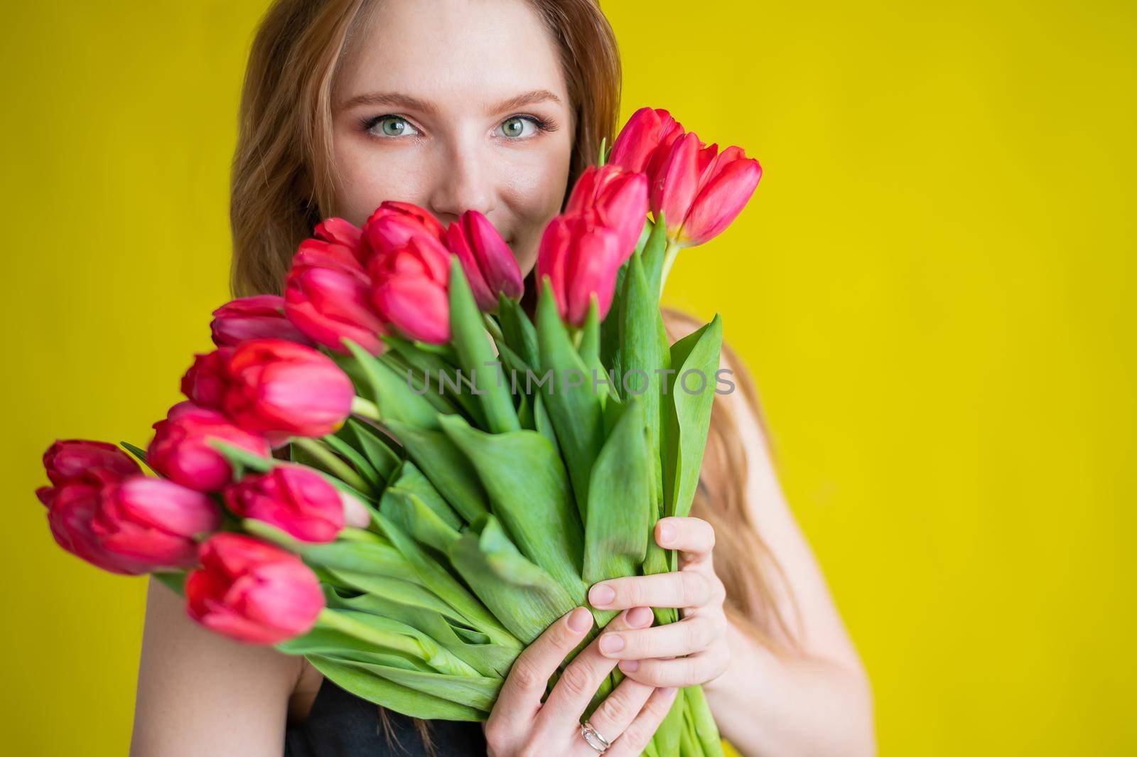 Woman with a bouquet of red tulips on a yellow background. Happy girl in a black dress holds an armful of flowers. Gift for Valentine's Day. The most romantic day of the year