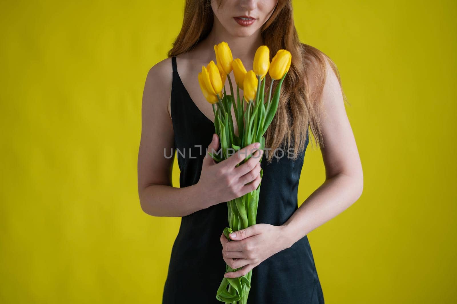 Unrecognizable woman with a bouquet of tulips on a yellow background. A girl in a black dress holds an armful of flowers. A gift for International Women's Day. Spring holiday. by mrwed54