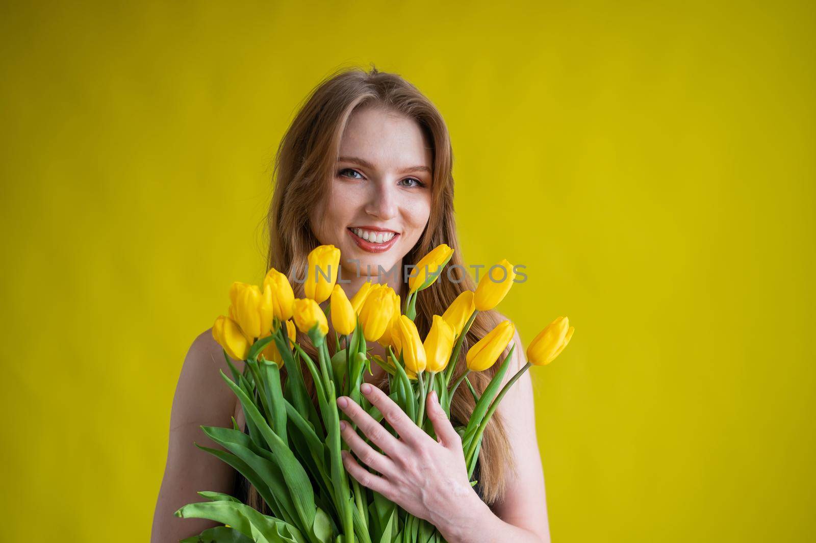 Caucasian woman with an armful of yellow tulips on a yellow background. International Women's Day. Bouquet of spring flowers.