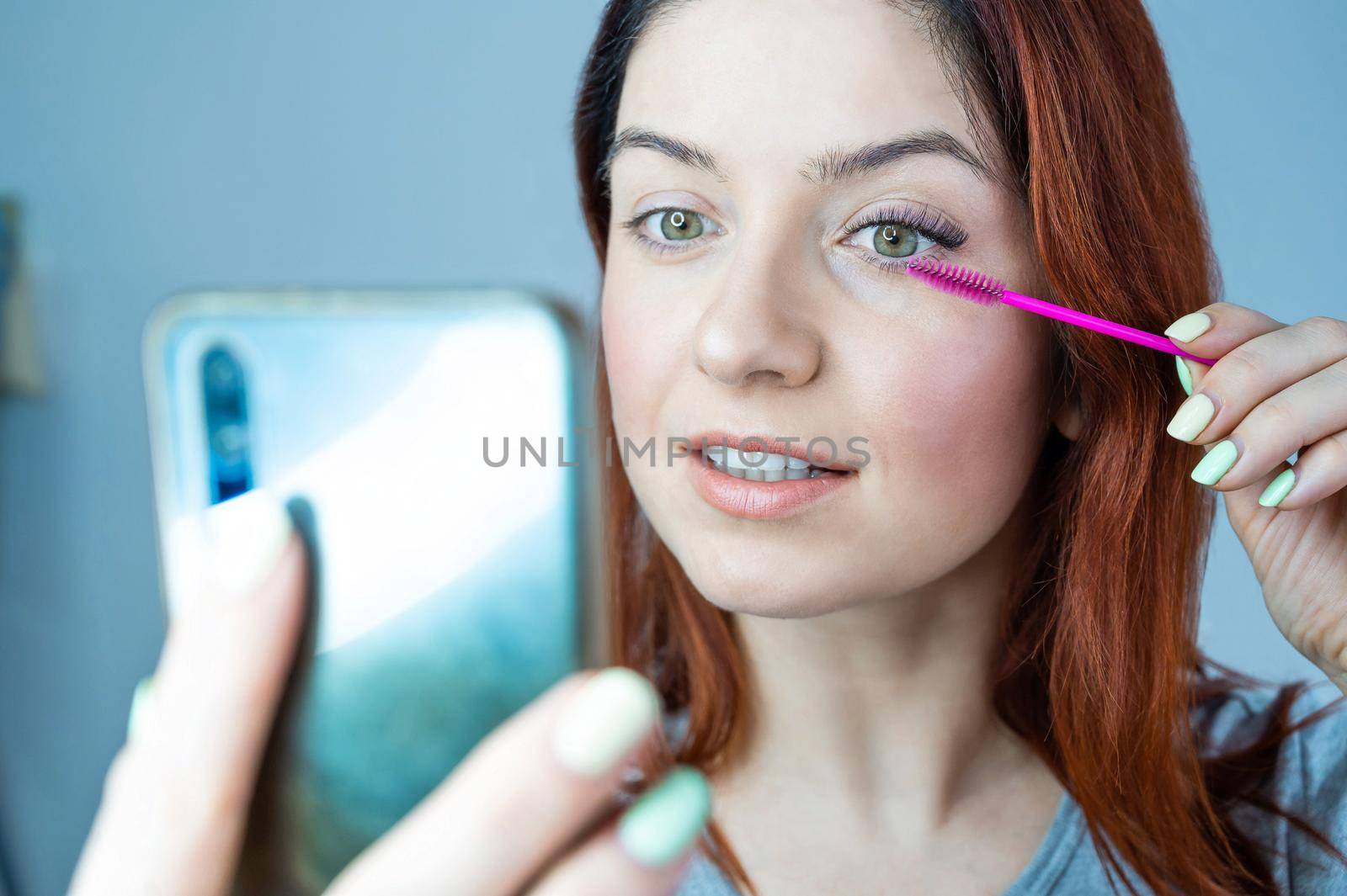 Beautiful redhead woman paints eyelashes and takes a selfie. The girl puts mascara and looks at the phone instead of a mirror