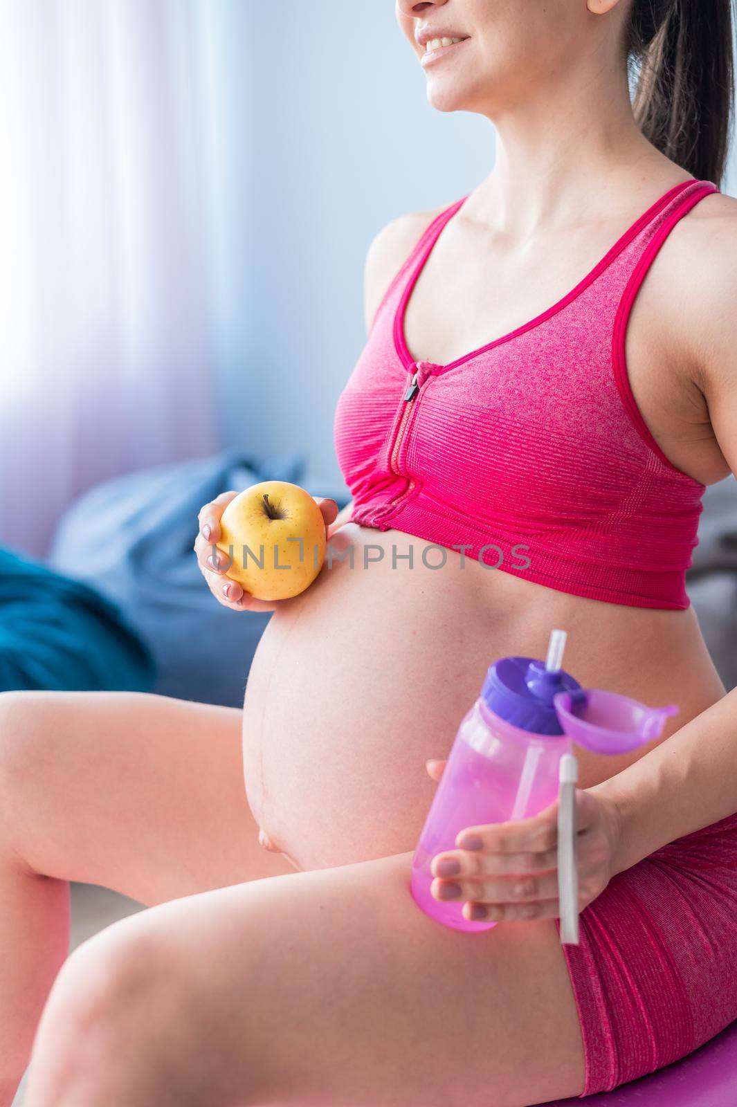 Faceless pregnant woman eating an apple after yoga. Expectant mother bites fruit and drinks water after Pilates. Girl sitting on a fitness ball