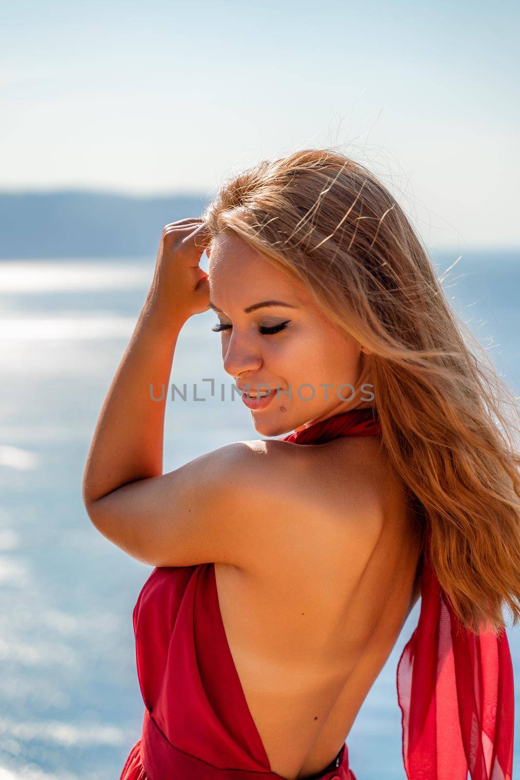 Smiling young woman in a red dress looks at the camera. A beautiful tanned girl enjoys her summer holidays at the sea. Portrait of a stylish carefree woman laughing at the ocean. by Matiunina