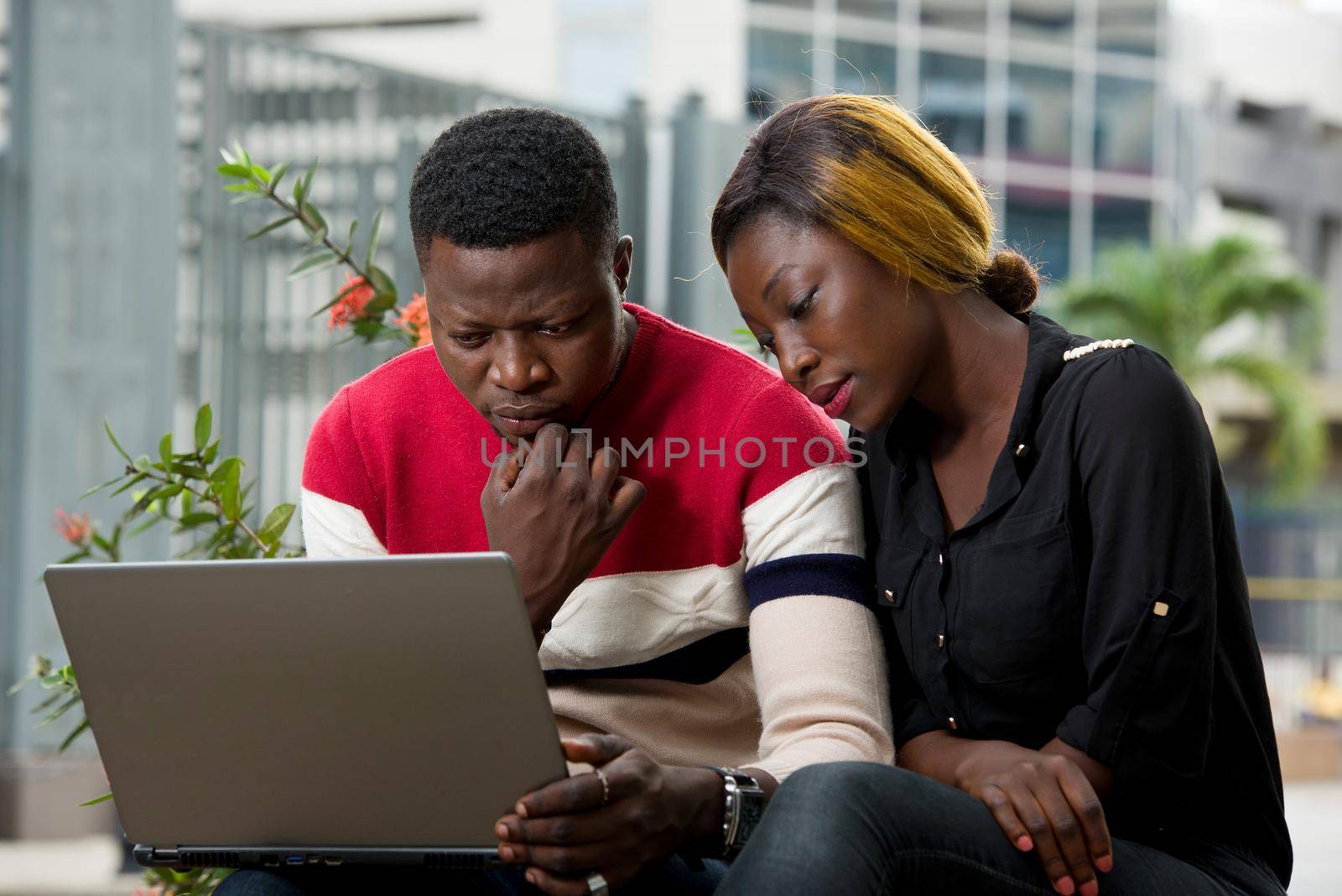 Two students sitting outside while working together on laptop on the side of the road outdoors in the summer..