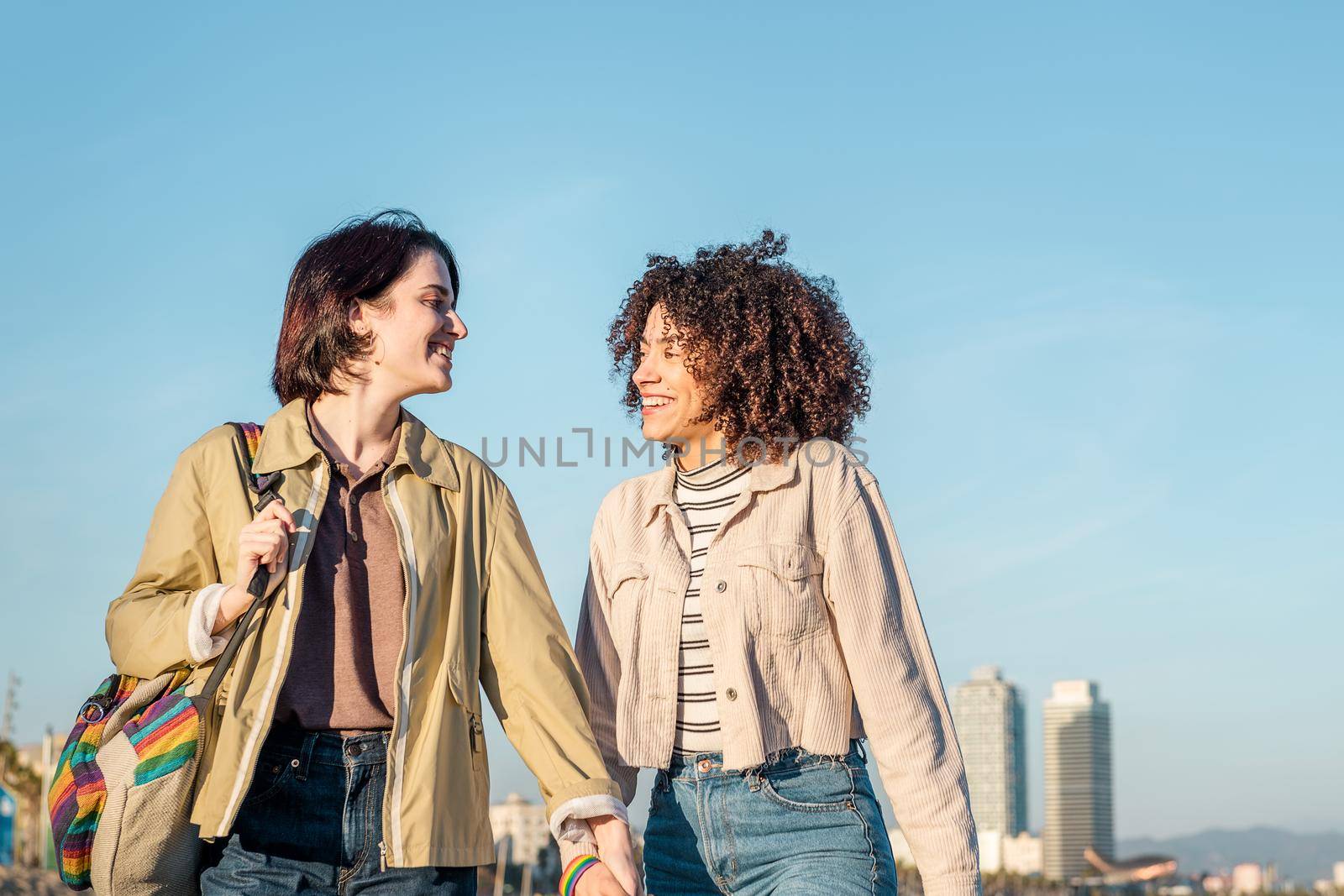 young lesbian couple walking holding hands and laughing in Barcelona, gay friendly city, with the colors of the rainbow flag, concept of sexual freedom and racial diversity