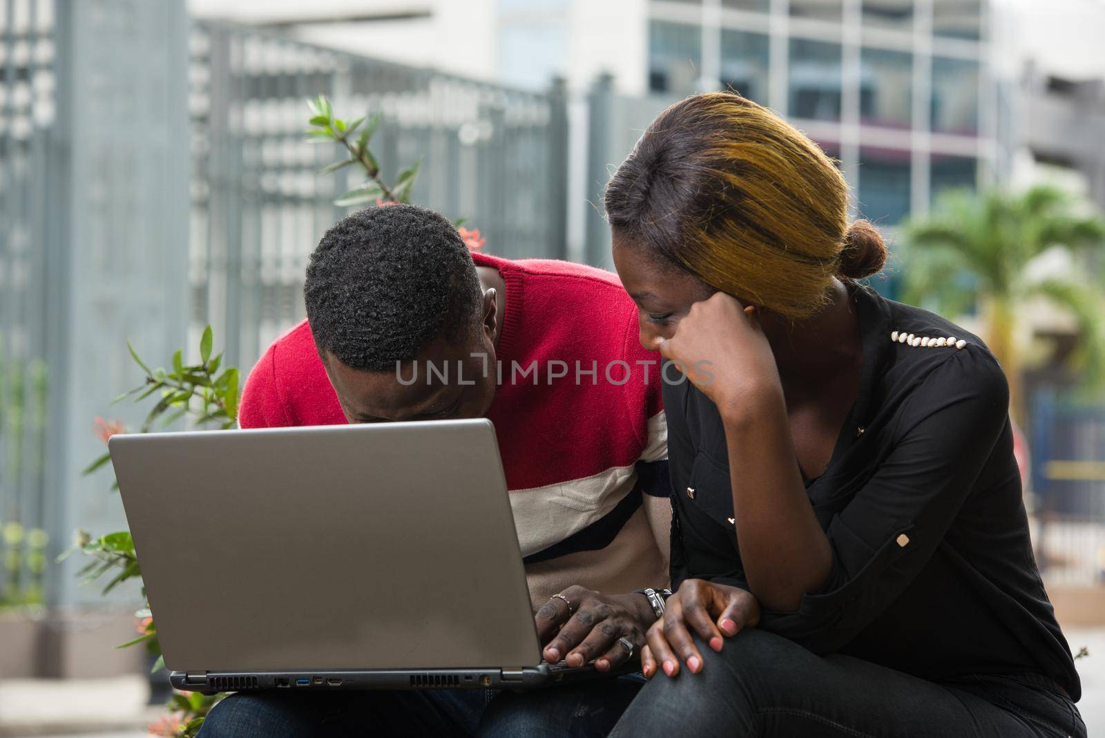 Two students sitting outside while working together on laptop on the side of the road outdoors in the summer.
