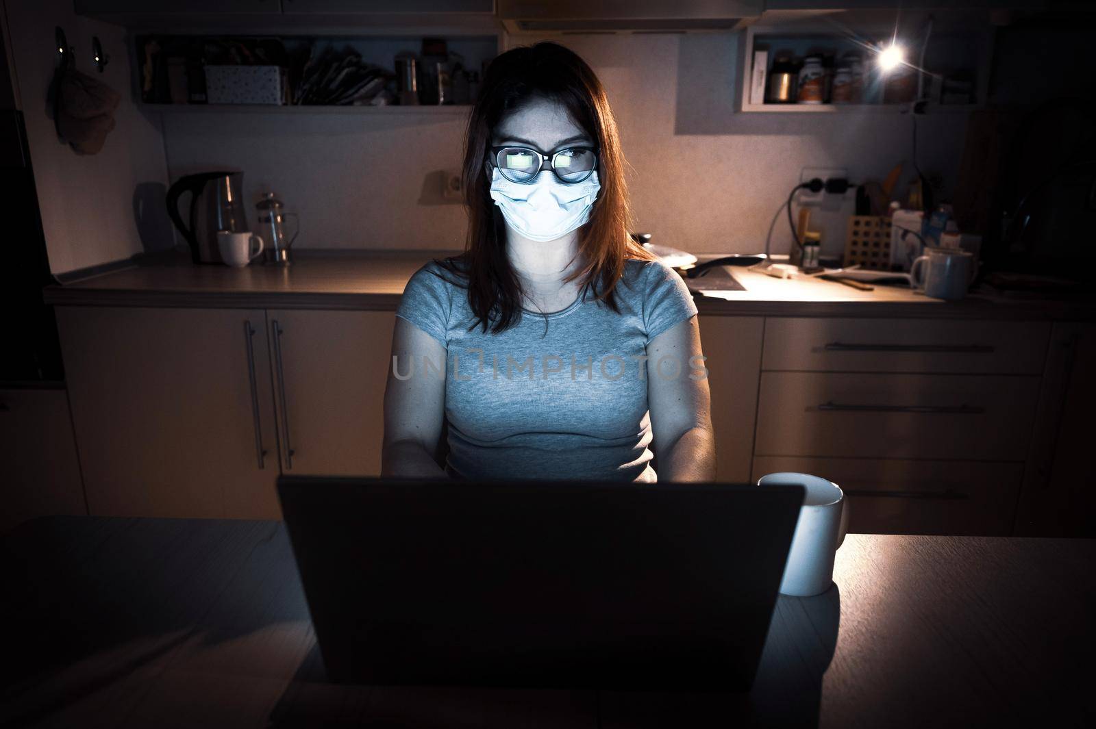 A girl in a mask and gloves learns during quarantine. A woman working overtime from home on a laptop. A freelancer is sitting in the kitchen in the dark in the light from the monitor