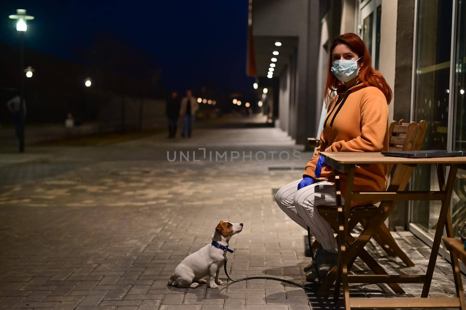 A woman in a medical mask and gloves walks late at night with her little dog. A girl sits on a chair in the street with a Jack Russell Terrier in quarantine. Coronavirus