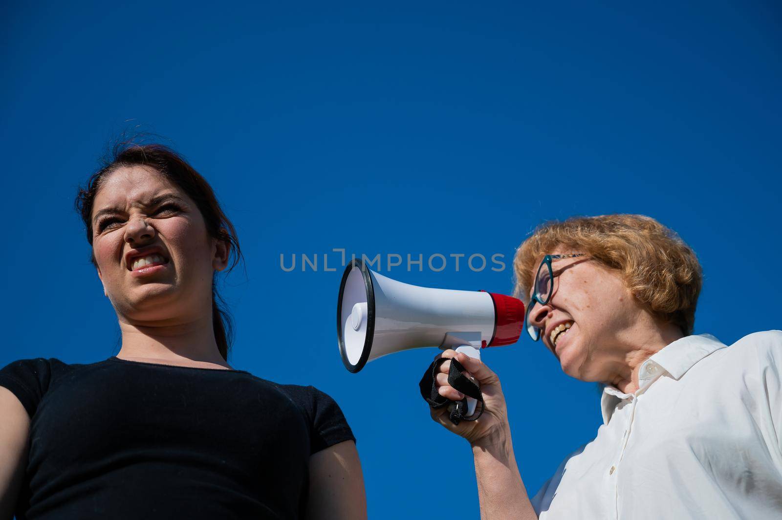 The conflict of generations. An emotional elderly woman shouting at her daughter in a megaphone. An elderly mother swears at a middle-aged woman on a loudspeaker on a blue background