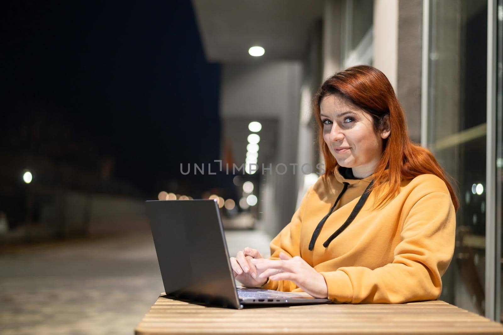 A woman works remotely at a laptop in a summer cafe late in the evening. A smiling girl is studying while sitting down in a wasted street at a wooden table