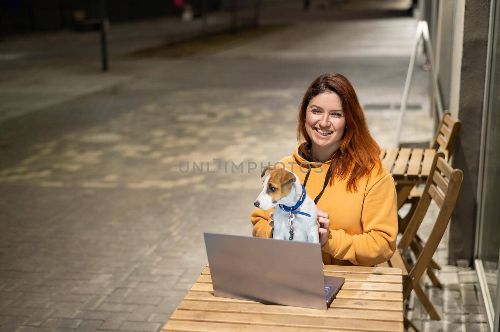 Smiling woman working on laptop at a wooden table in the street. The girl looks at the monitor with her pet jack russell terrier. Freelancer walks the dog in the evening. by mrwed54