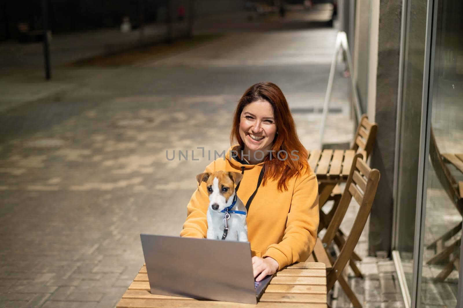 Smiling woman working on laptop at a wooden table in the street. The girl looks at the monitor with her pet jack russell terrier. Freelancer walks the dog in the evening