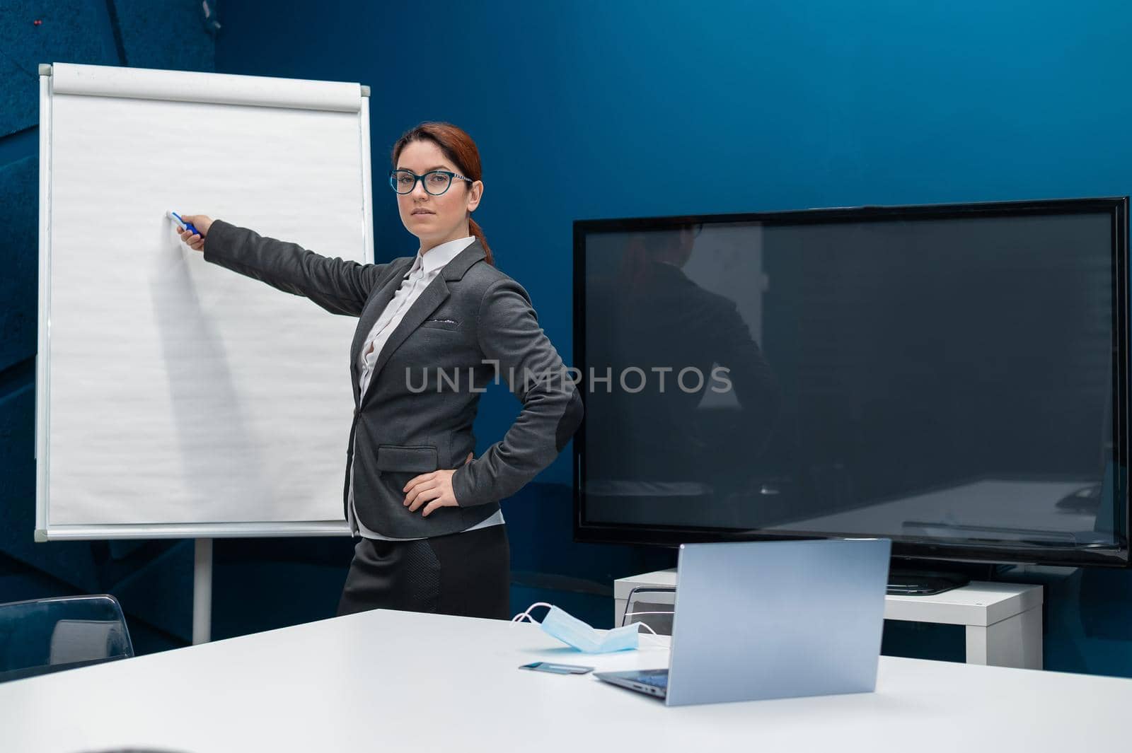 Friendly woman in a suit writes on a blank white board with a marker. Red haired girl makes a presentation in the office. Beautiful female business coach at a conference