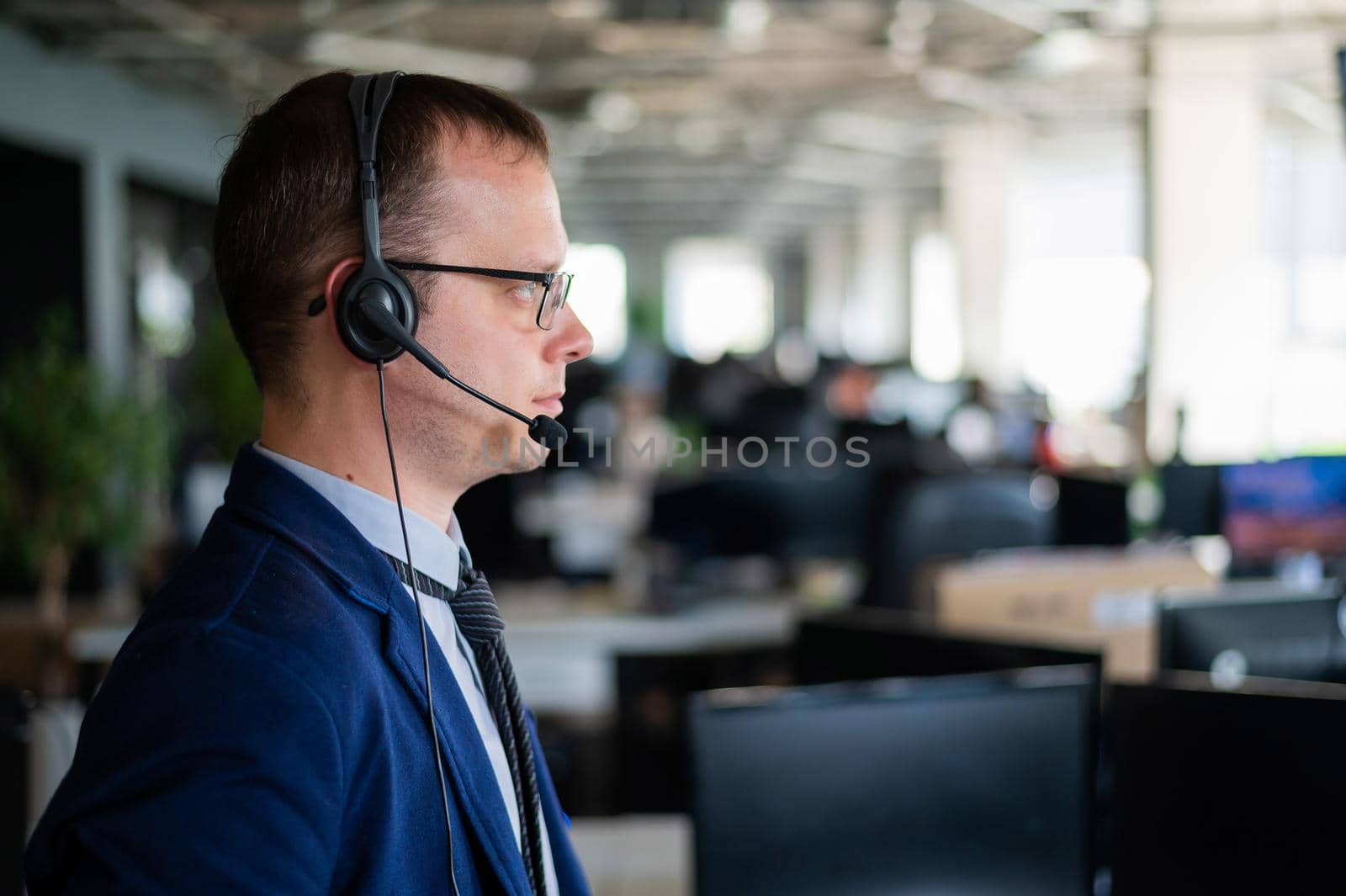 Portrait of a male call center operator in headset at workplace. A man works in an open space office answering customer calls. Support service or hotline. Agent of a telemarketing company