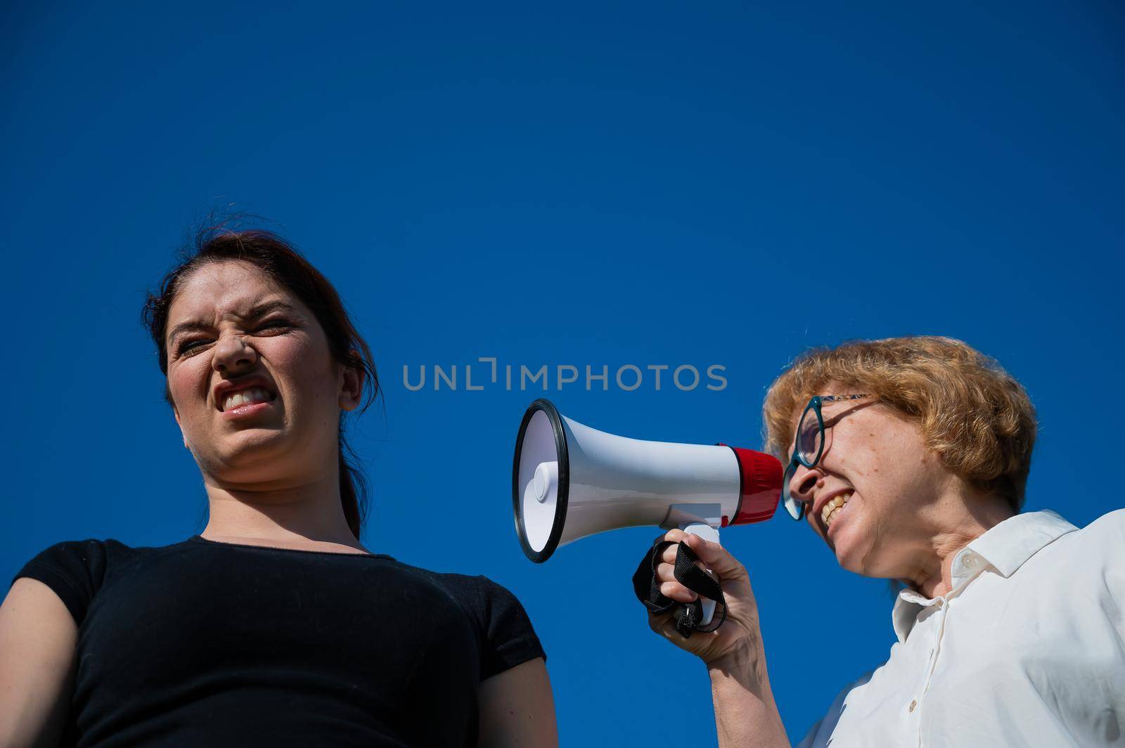The conflict of generations. An emotional elderly woman shouting at her daughter in a megaphone. An elderly mother swears at a middle-aged woman on a loudspeaker on a blue background