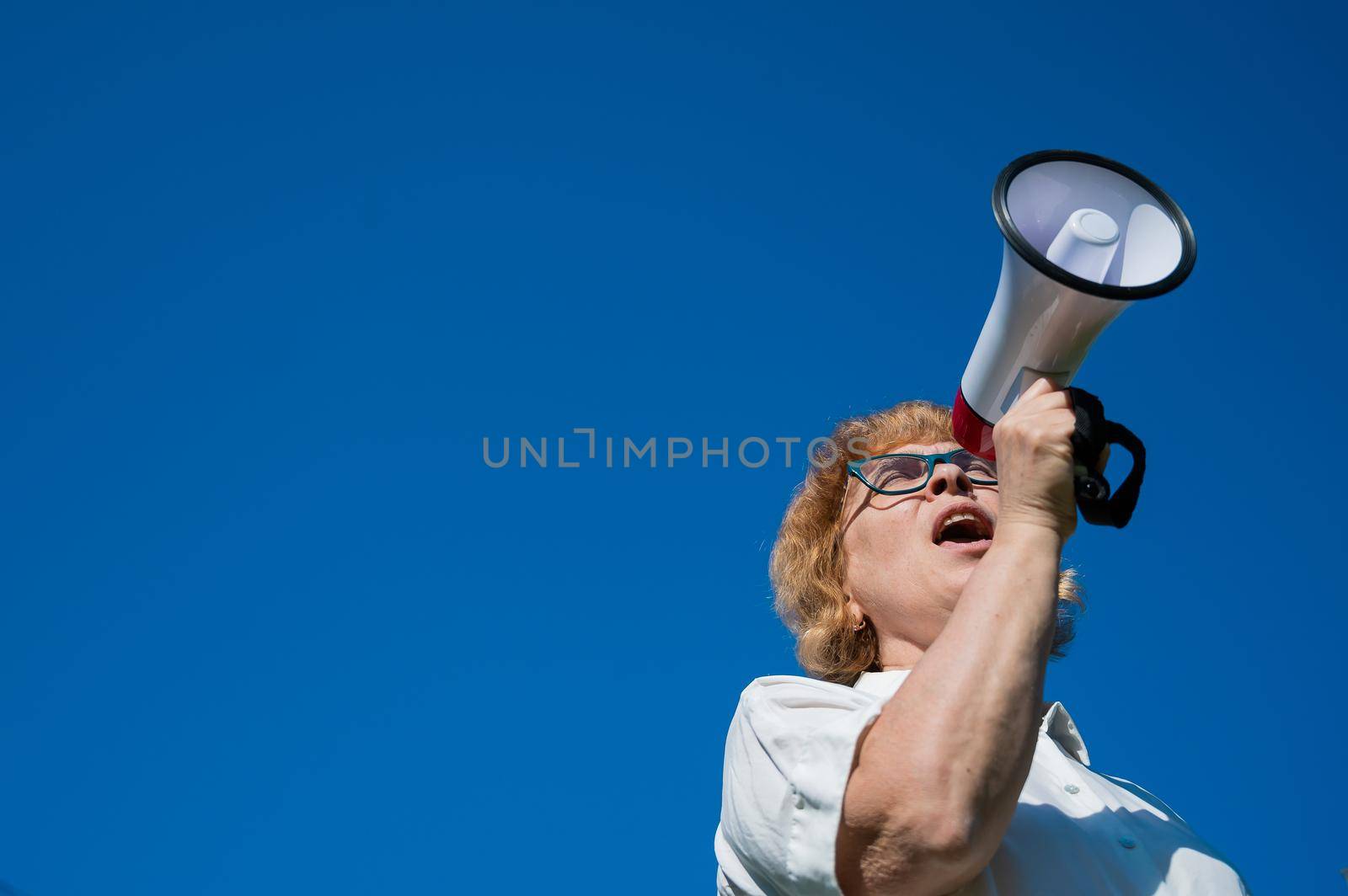 Emotional senior woman makes a speech in a megaphone on the outside. A pensioner yells into a sound amplifier on a blue background. female leader of the rally voices the conditions in the loudspeaker. by mrwed54