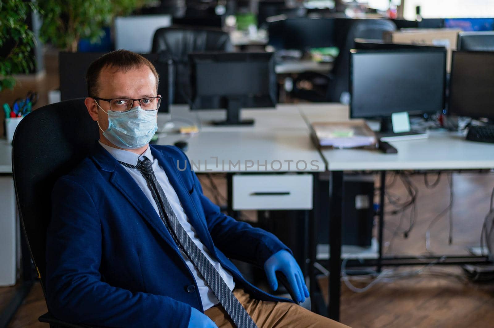 Serious man in a business suit and glasses in an empty office during quarantine. Male manager in a medical mask at the workplace. Social distance and workspace disinfection. by mrwed54