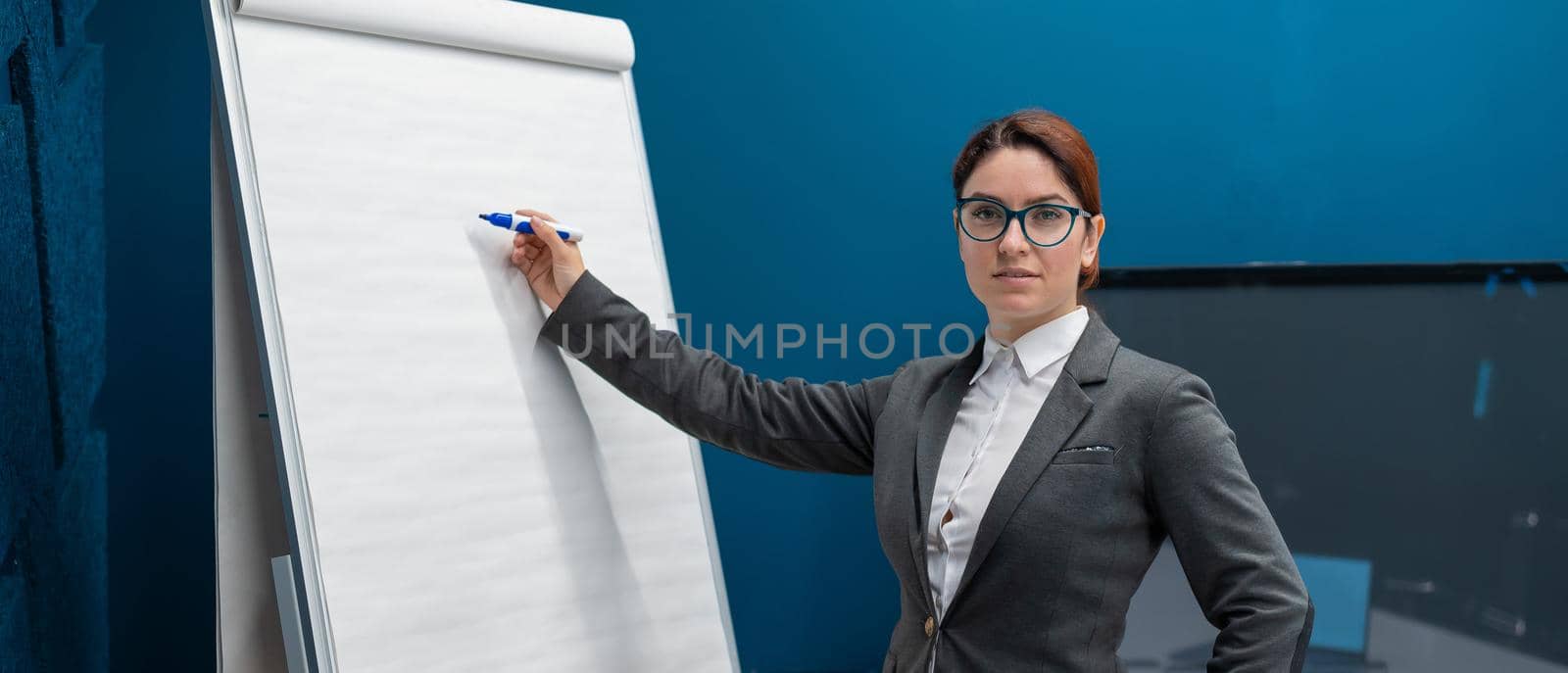 Friendly woman in a suit writes on a blank white board with a marker. Red-haired girl makes a presentation in the office. Beautiful female business coach at a conference. by mrwed54