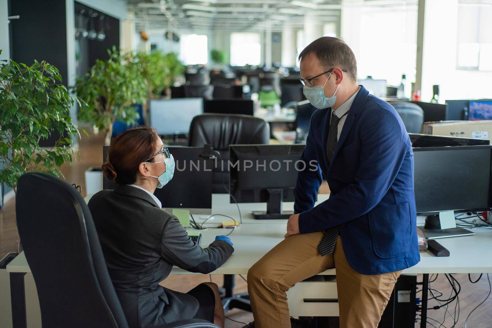 Colleagues work in medical masks in an open office space communicate at the desk. A man and a woman in office suits are discussing a working draft. The boss controls the subordinate