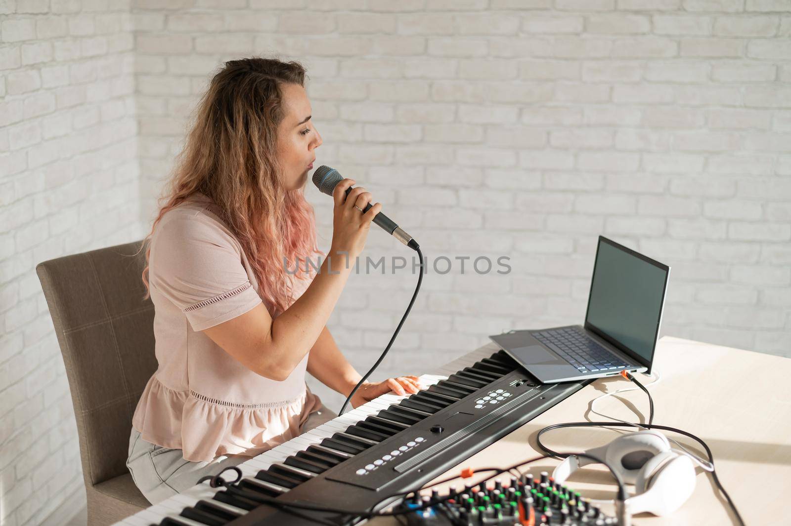 A woman records a vocal lesson using a laptop and accompanying on a keyboard while at home. The teacher sings a song into the microphone and plays the electronic piano. A blogger is recording a video