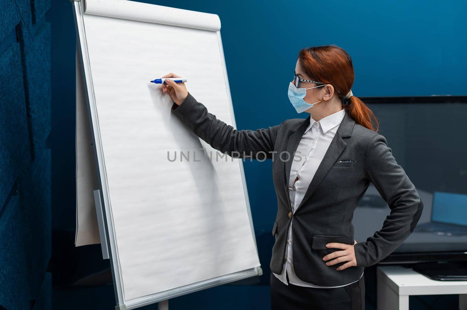 A woman in a medical mask leads a seminar in a conference hall. Business coach writes on a paper white board in the office. Female employee demonstrates presentation while maintaining social distance. by mrwed54