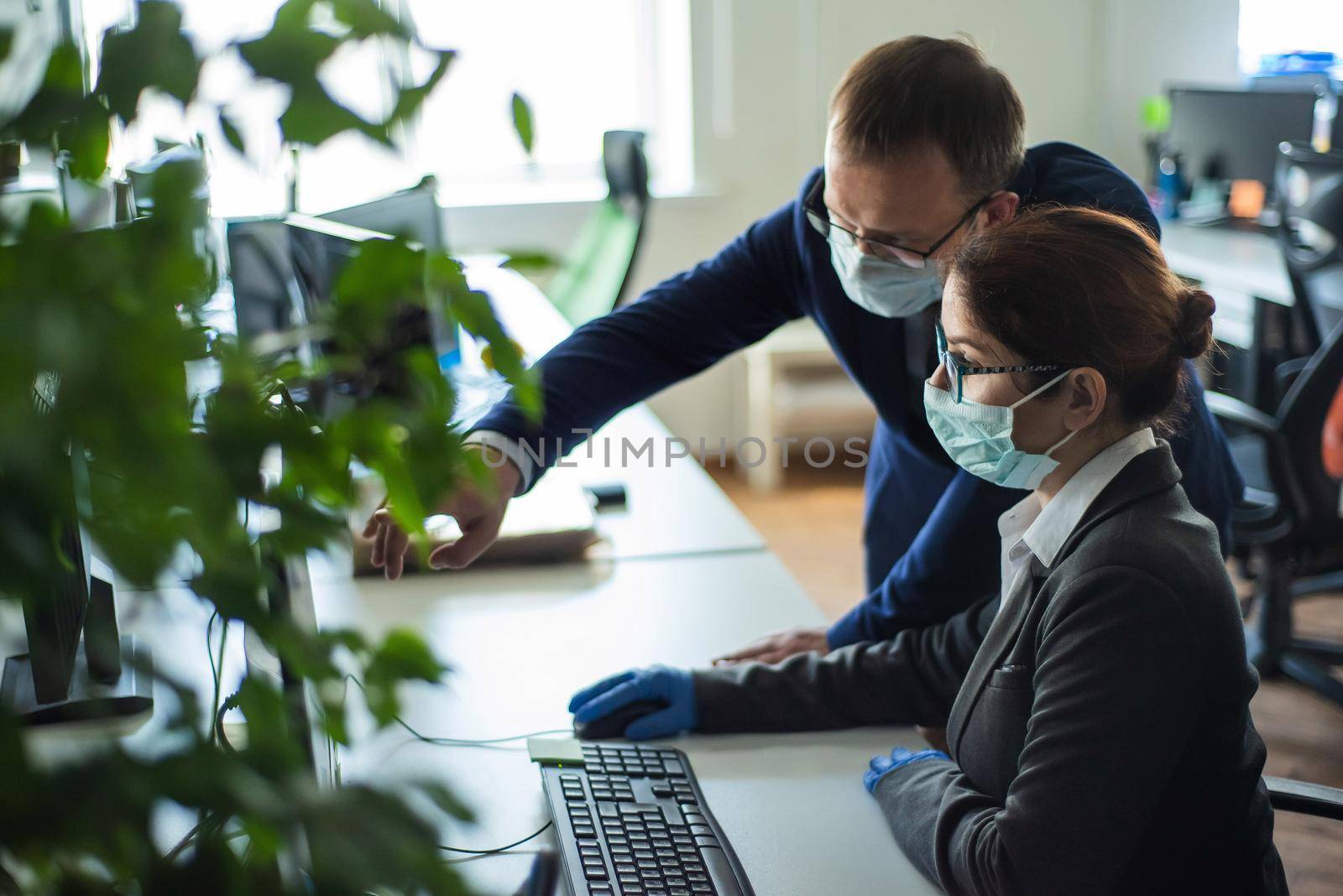 Colleagues in surgical masks in an open office space communicate at the work desk. A male top manager teaches a female intern how to work at a computer. The boss directs the subordinate. by mrwed54