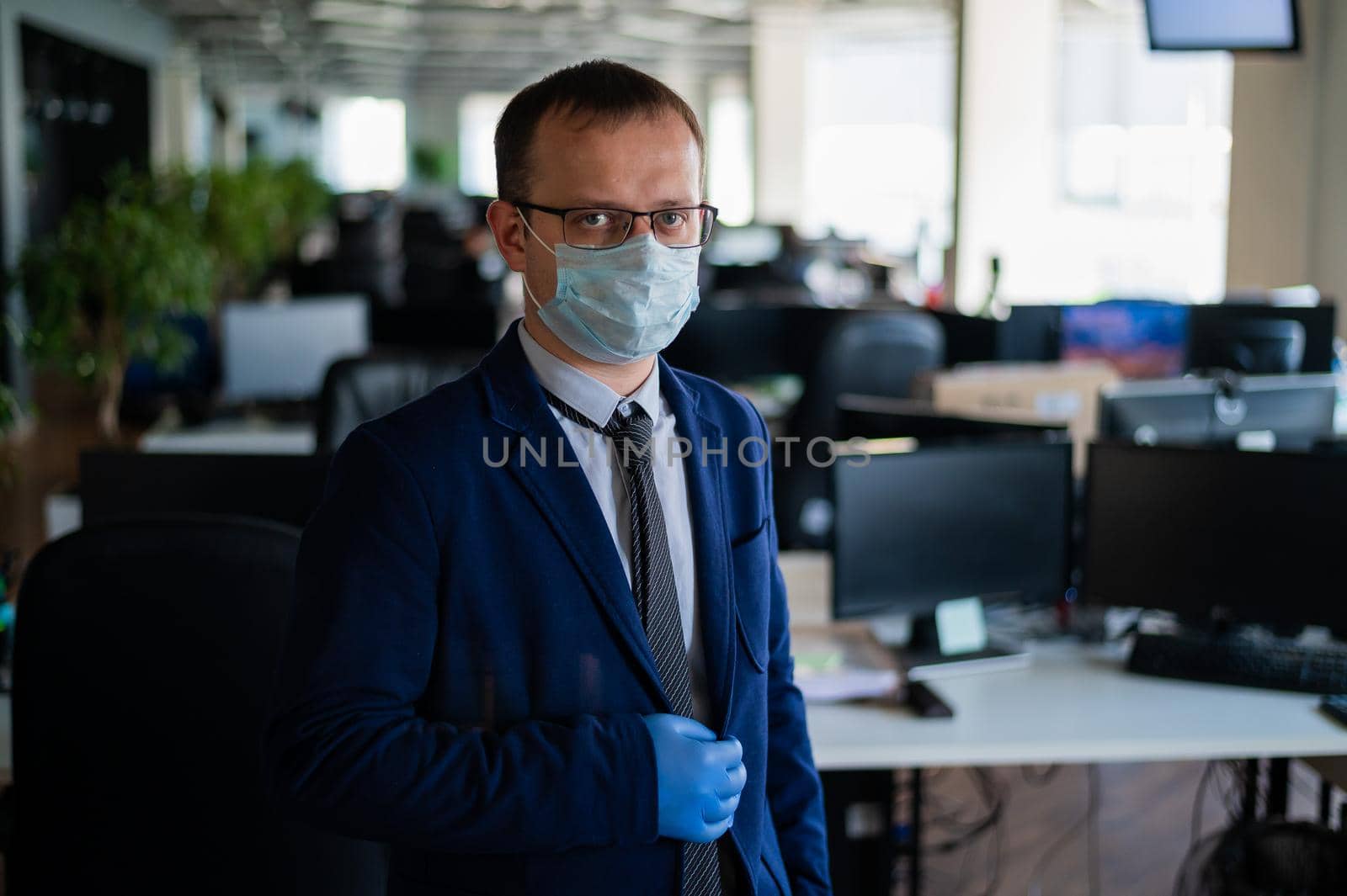 Serious man in a business suit and glasses in an empty office during quarantine. Male manager in a medical mask at the workplace. Social distance and workspace disinfection