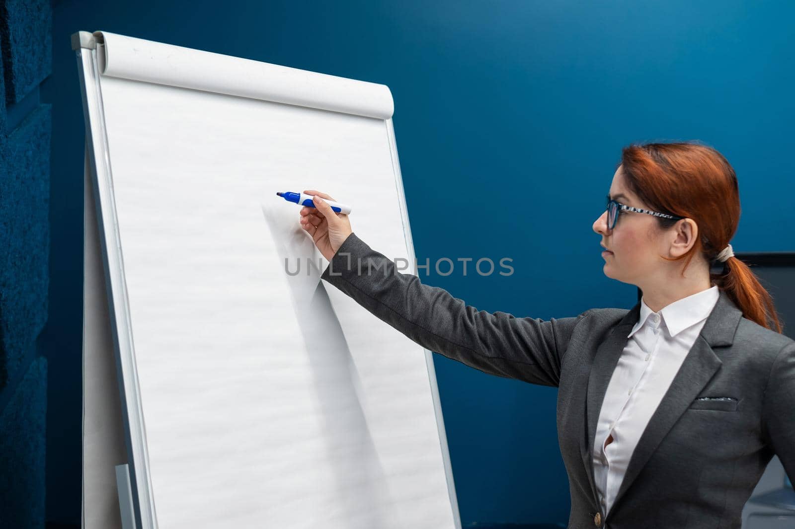 Friendly woman in a business suit writes on a blank white board with marker. Red-haired girl makes a presentation in the office. Beautiful female teacher in the classroom