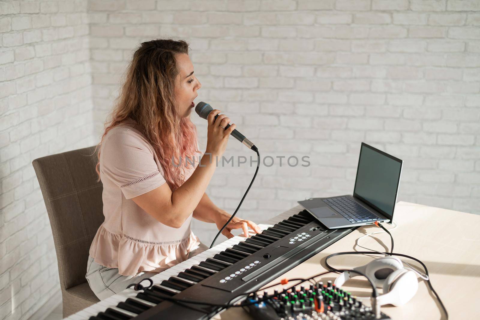 A woman records a vocal lesson using a laptop and accompanying on a keyboard while at home. The teacher sings a song into the microphone and plays the electronic piano. A blogger is recording a video