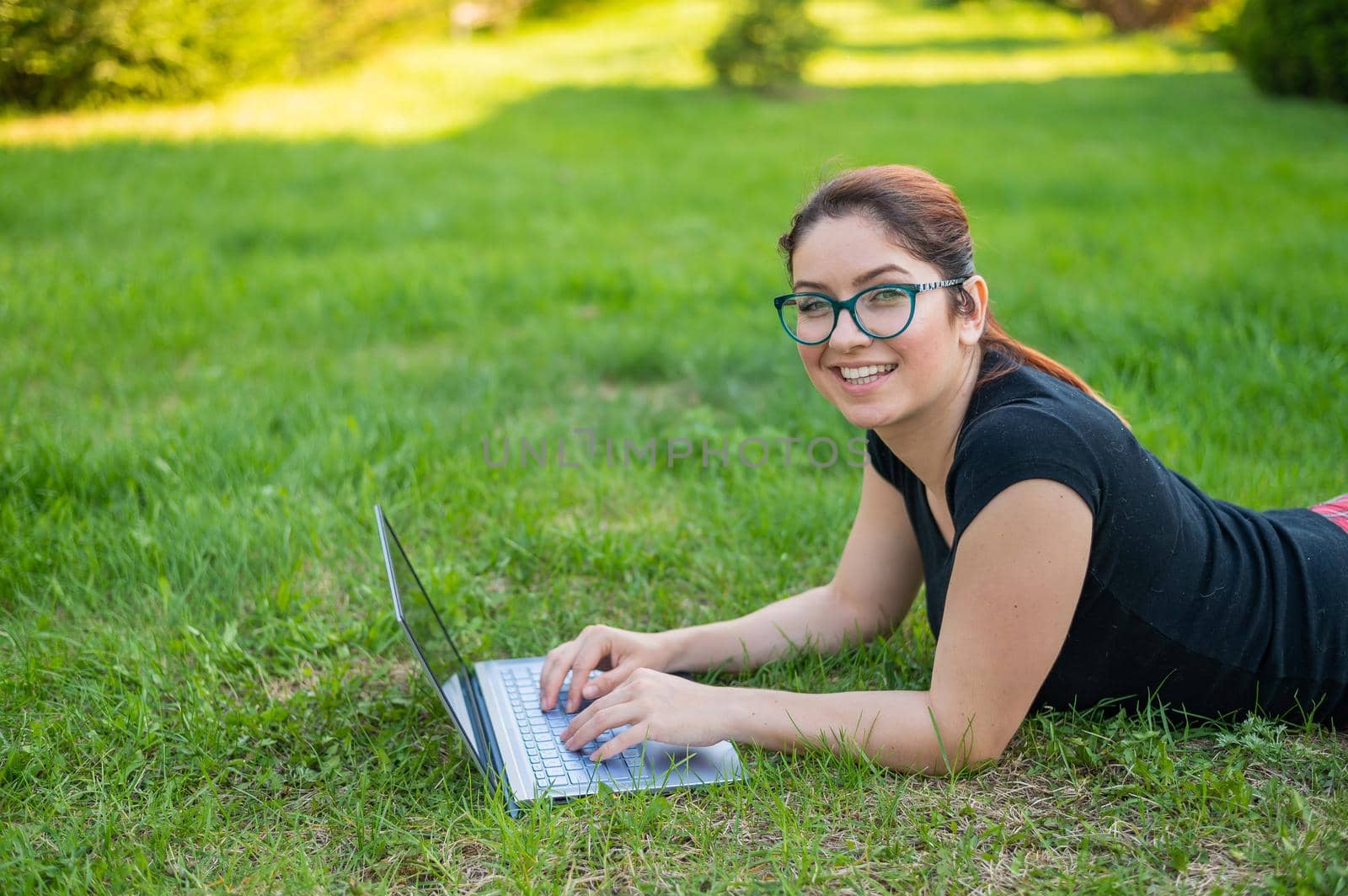 Happy red-haired girl in glasses lies on the lawn in the park and types on the laptop keyboard. Young caucasian female freelancer works remotely on a wireless computer outdoors. by mrwed54