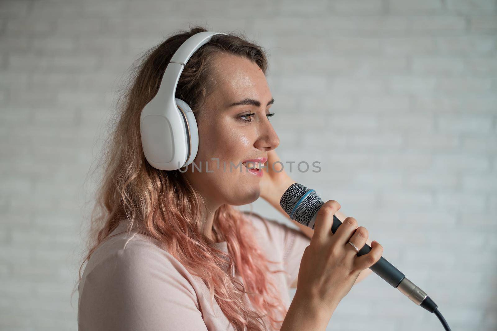 Close-up portrait of a caucasian woman with curly hair singing into a microphone. Beautiful sensual girl in white headphones sings a song in home karaoke