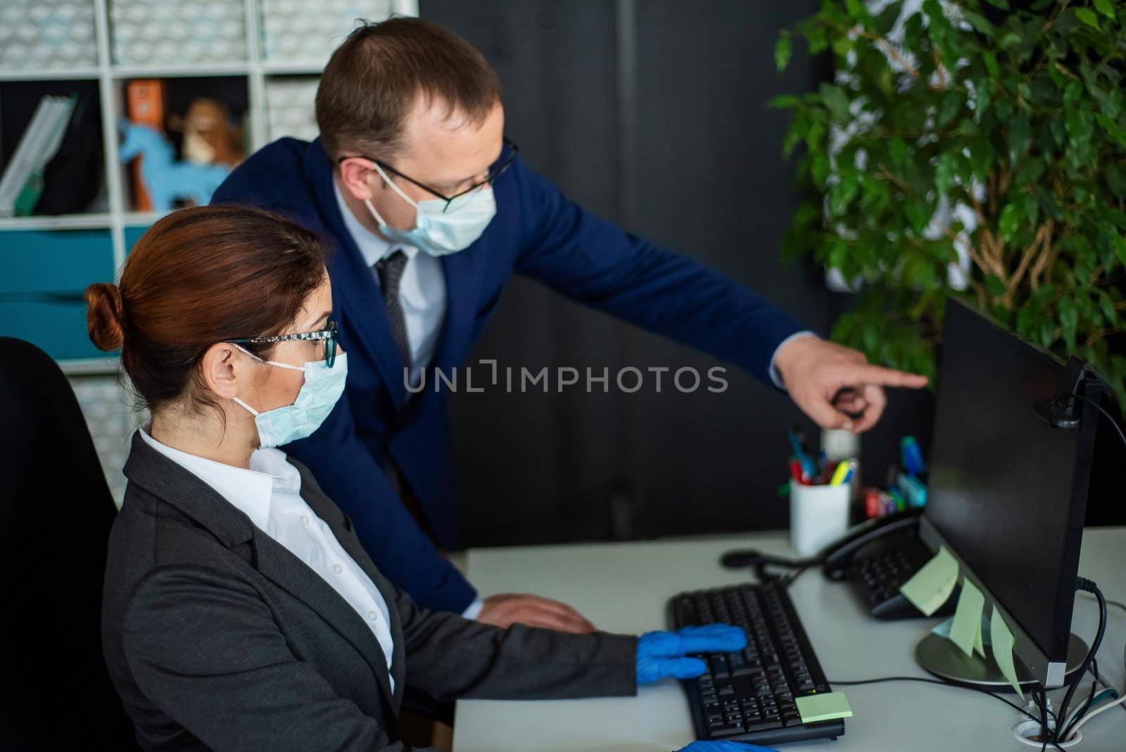 Colleagues in surgical masks in an open office space communicate at the work desk. A male top manager teaches a female intern how to work at a computer. The boss directs the subordinate. by mrwed54