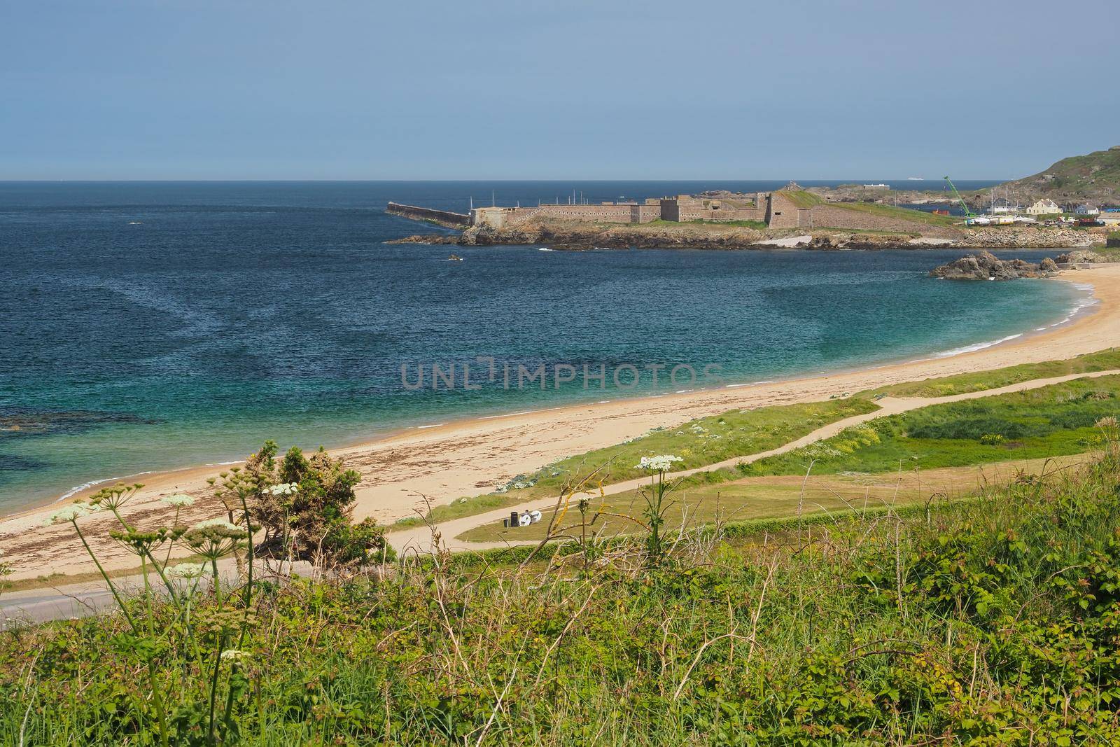 View across bay and sandy beach to Fort Grosnez and Braye Harbour from Fort Tourgis under blue sky, Alderney, Channel Islands