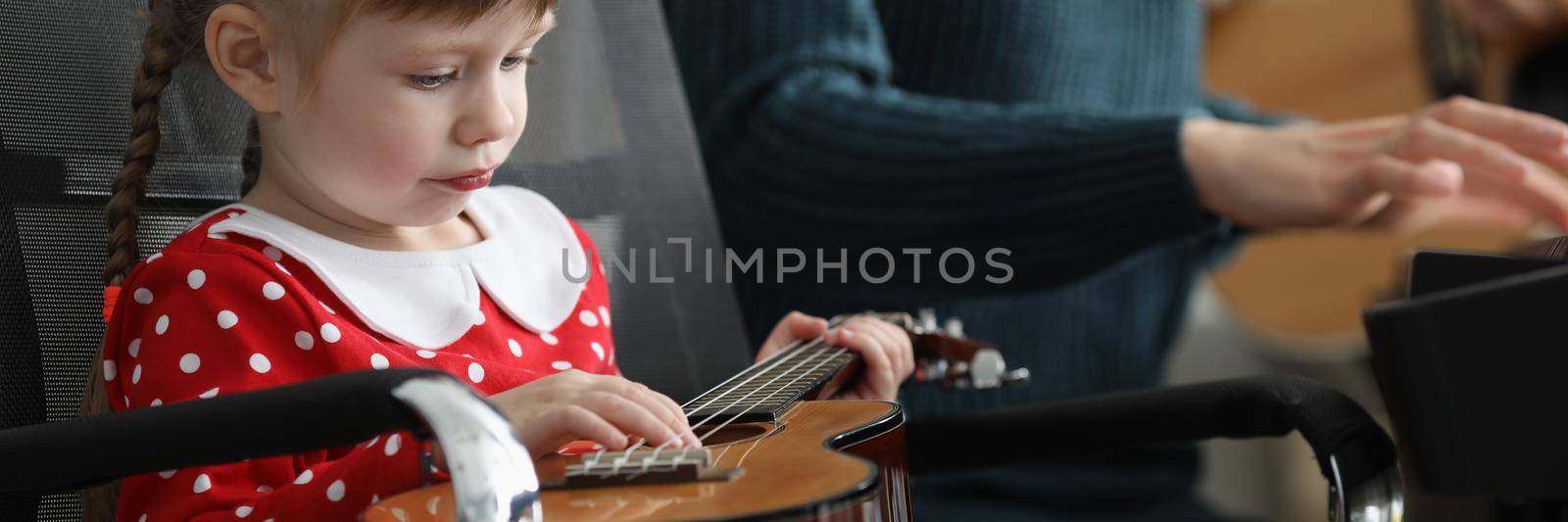 Portrait of bored girl hold guitar and fooling around with musical instrument. Child pull string while teacher play piano. Preschool development concept