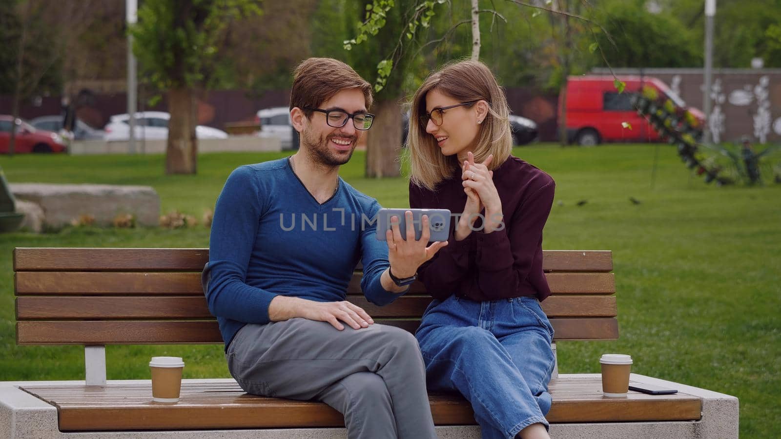 Young smiling couple conducting a video call on phone. Pretty Caucasian woman and man sitting outdoor beach coffee break. Phone video call conversation.
