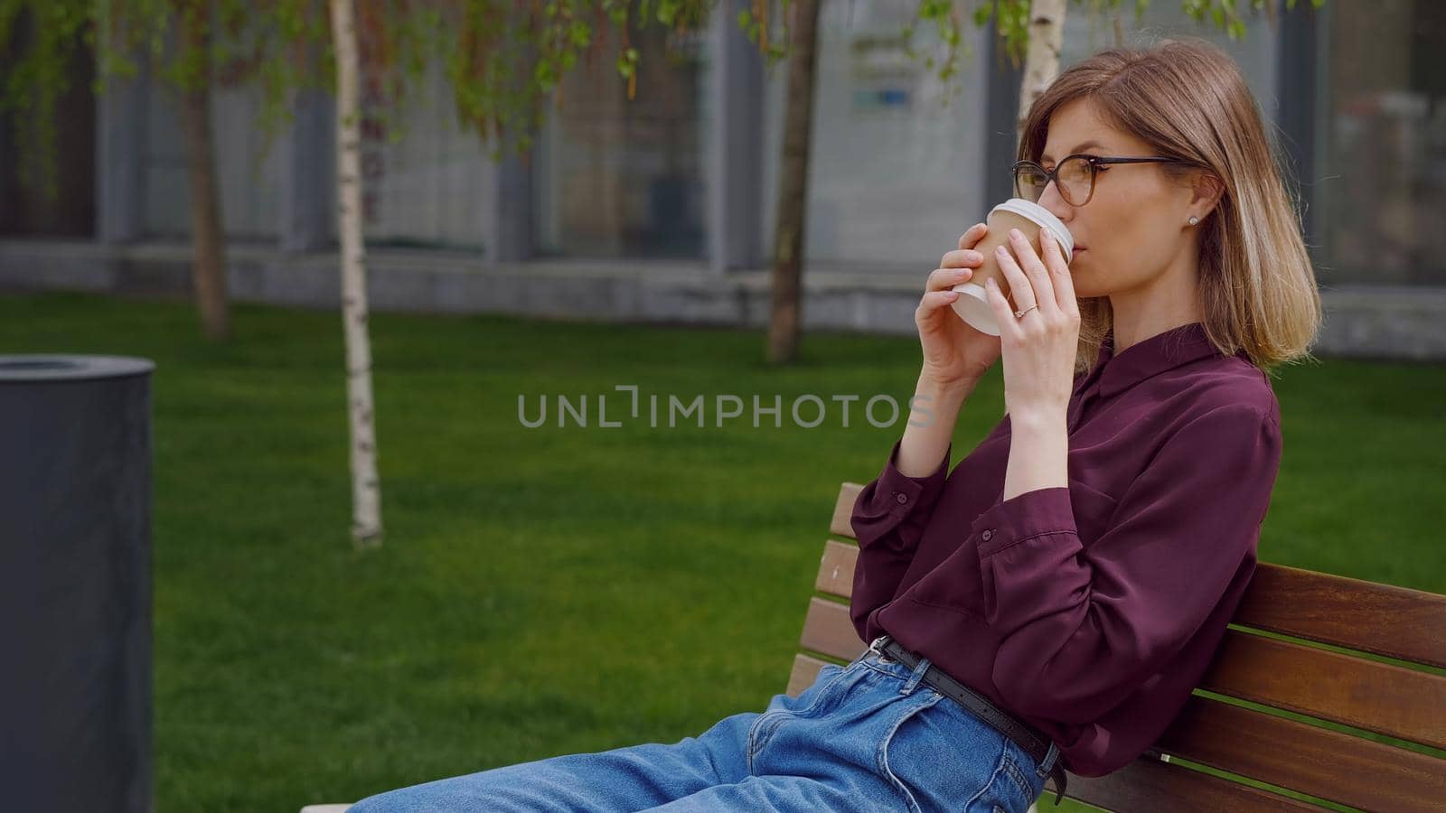 Businesswoman takes a break from work and sits on a bench outside her office while sipping coffee to-go. Wearing casual style. Sunny day. Portrait pretty successful Caucasian lady. Slow motion video.