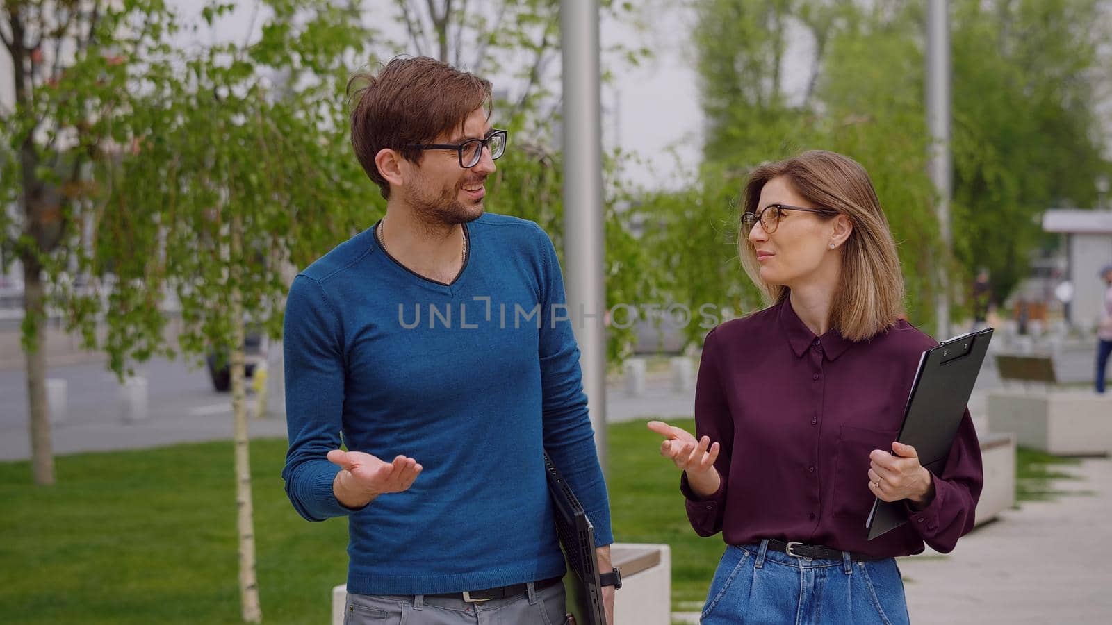 Man and woman walk outside talking about business. Concept between partners. Man holds the laptop in his hand, the woman has a clipboard. Successful people. All about job. Summer. Slow motion video.