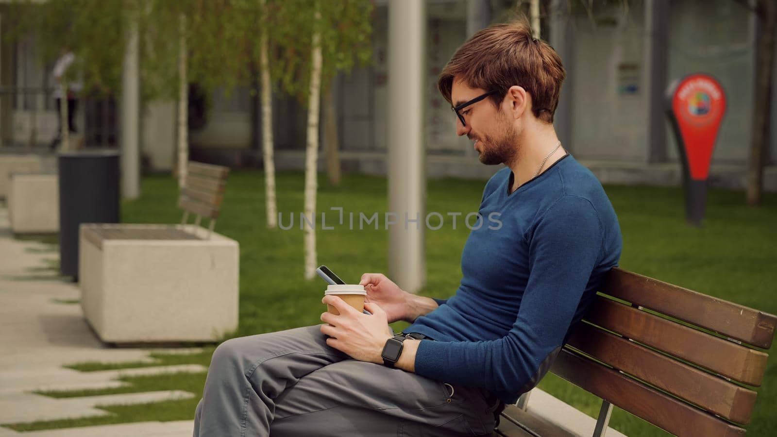 Outdoor scene of handsome young man browsing smartphone social network app while drink coffee. Student man relaxing in a park.