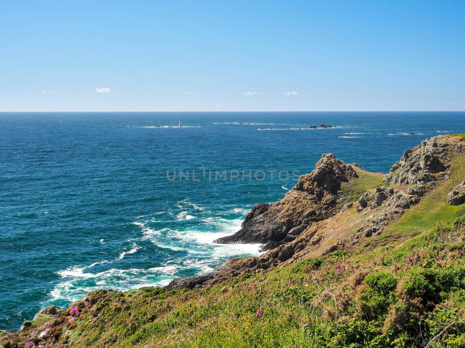 View across rocky bay to Les Hanois Lighthouse on Le Biseau rock, under blue sky and white clouds, Pleinmont, Guernsey, Channel Islands