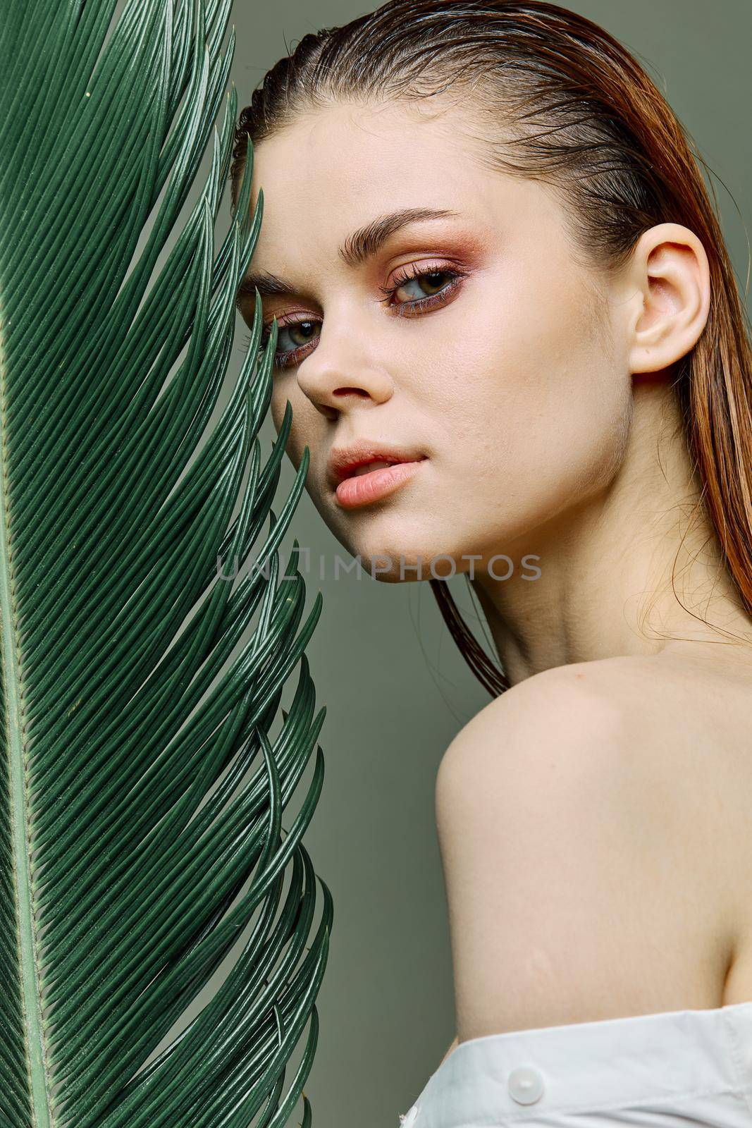 a sophisticated, elegant woman with evening makeup, stands with a green palm leaf, covering part of her face with it. Close vertical portrait, photo without retouching. High quality photo