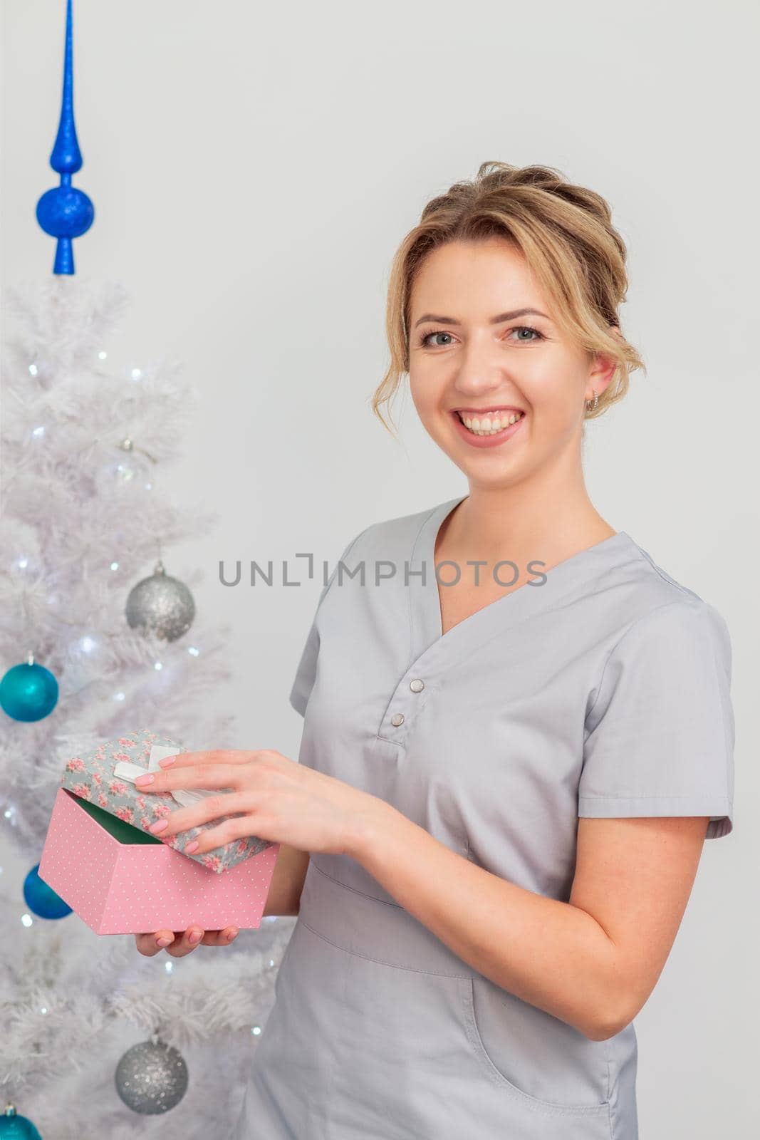 Beautiful young caucasian woman smiling in medical uniform with gift box near christmas tree on light background
