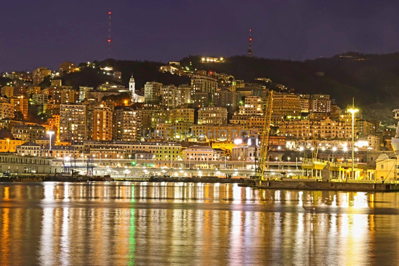 Genova, Italy - February 22, 2022: Evening view of Genoa (Genova) port, Italy with port cranes and industrial zone. Genoa, Italy. Lanterna lights by night. City illuminated behind the sea.