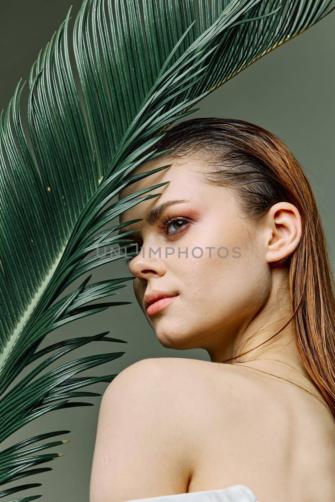 a sophisticated, elegant woman with evening makeup, stands with a green palm leaf, covering part of her face with it, looking away. Vertical photo without retouching by Vichizh