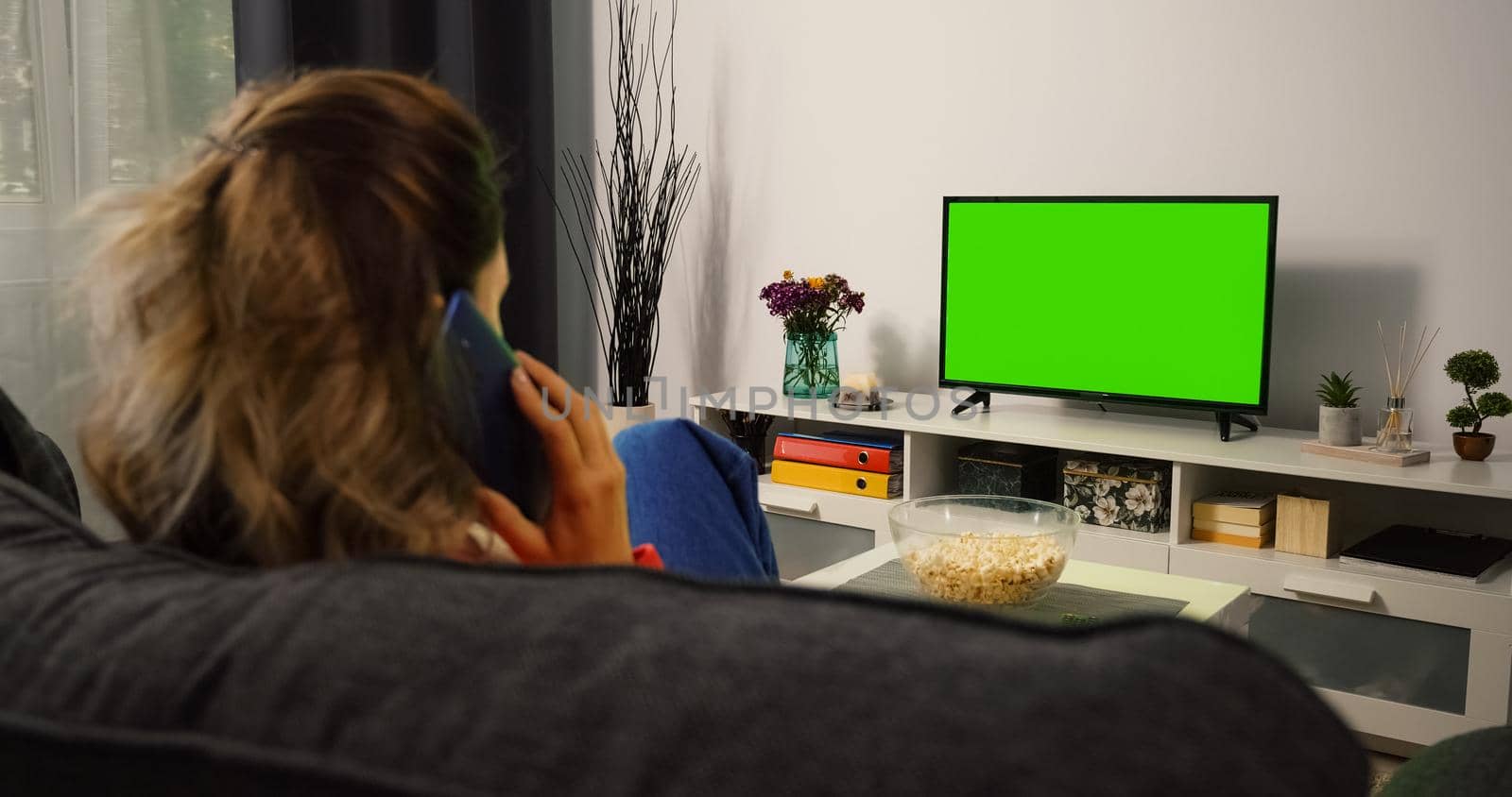 Woman watching Tv Green Chroma Key Screen and talking on Phone. Young woman have a phone conversation while looking Tv in the evening. Back view watching Green Screen.