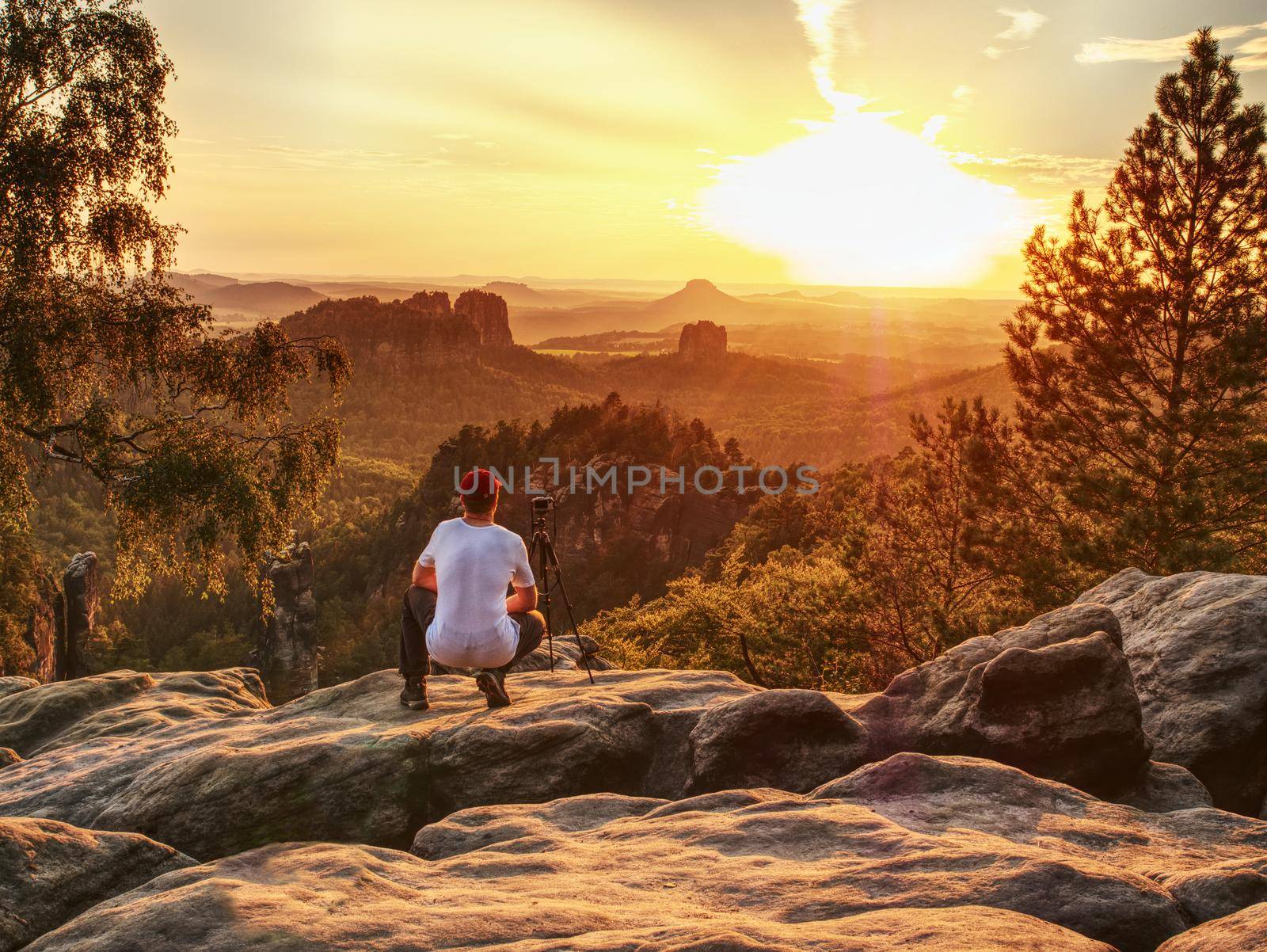 Nature photographer takes photos on peak of rock. A mirror camera and tripod. Dreamy fogy landscape, spring orange pink misty sunrise in beautiful valley below.