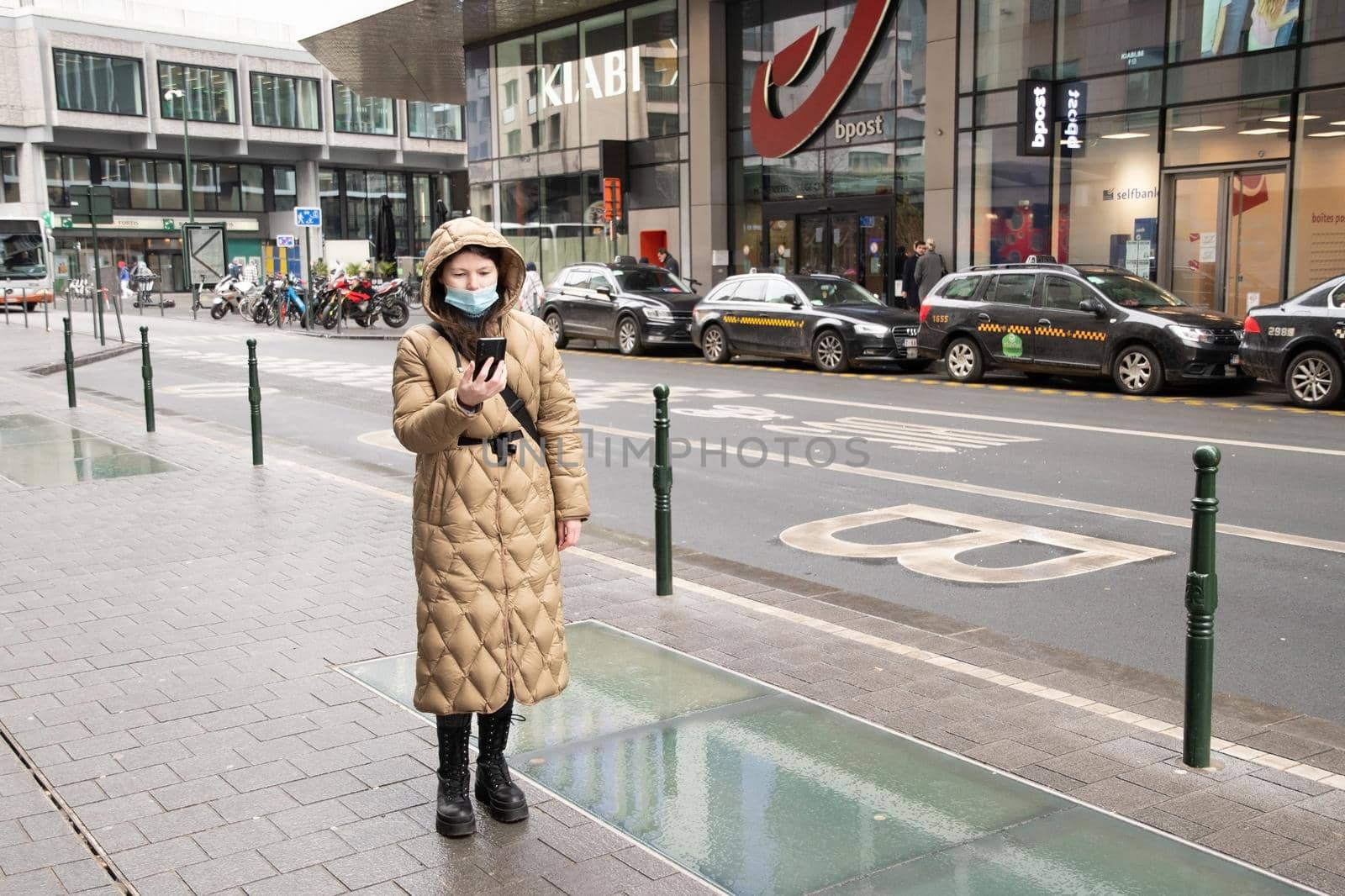 Brussels Belgium - 01.31.2021 young woman in a warm down jacket in a hood and a mask walks down the street of Brussels in rainy weather. High quality photo